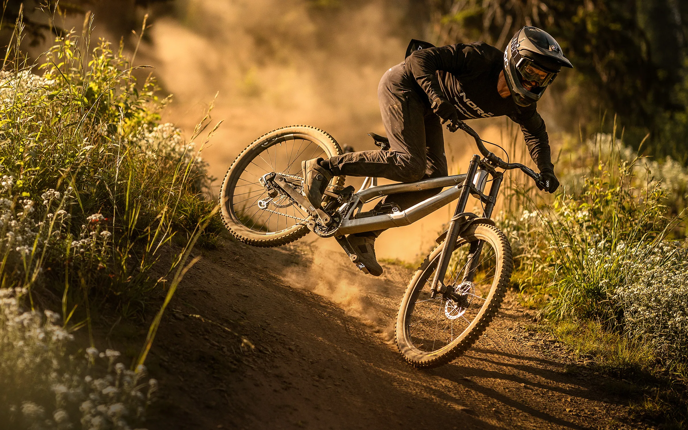 Norco downhill rider Kirk McDowall leans hard into a dusty berm, backlit by golden light and framed by wildflowers.